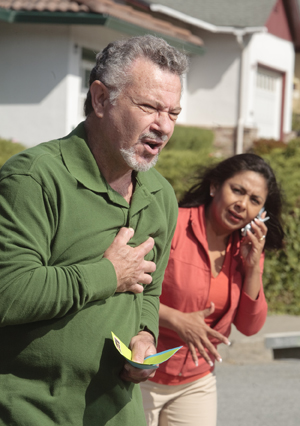 Man holding hand to chest in pain as worried woman next to him calls for help on cell phone.