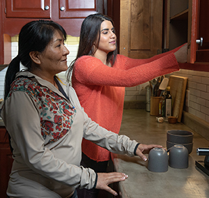 Woman getting dishes from cupboard with younger woman helping.