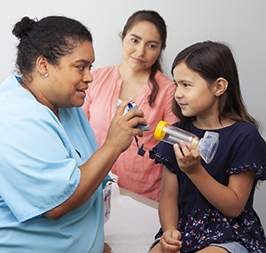 Una proveedora de atención médica le muestra a una niña cómo usar un inhalador de dosis medida con un espaciador y una máscara. Mujer parada cerca.